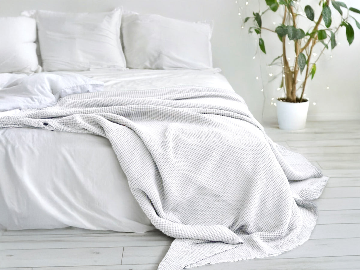 In einem Schlafzimmer liegt eine Gletscher-weisse und eine Nebel-graue Tagesdecke aus der beties "PUR.NATURE" Serie auf einem Bett ausgebreitet.