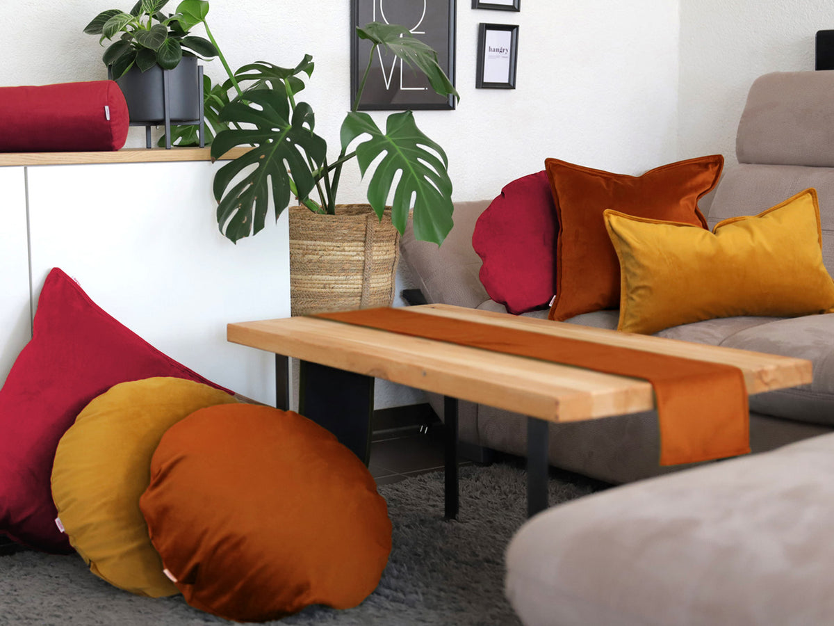 Karmin-rote, rost-orangene und curry-senf-gelbe Samt Kissenbezuege und ein rost-oranges Samt Tischband sind in einem Wohnzimmer dekoriert.