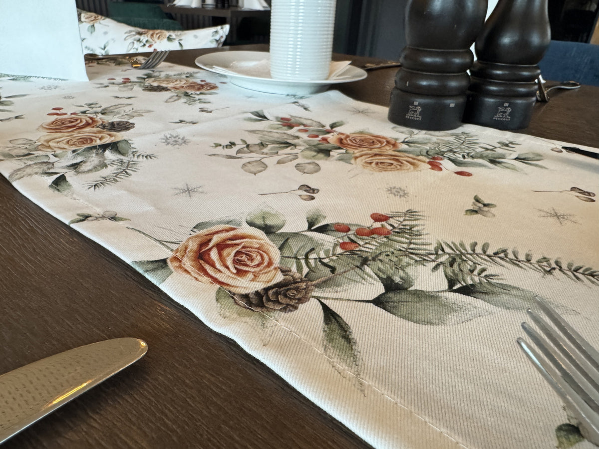 Weisser Tischlaeufer mit gefrosteten Rosen in orange, Kieferzapfen und Zweigen aus der beties Magie des Winters Kollektion "Frost-Rose" auf einem gedeckten Holztisch.