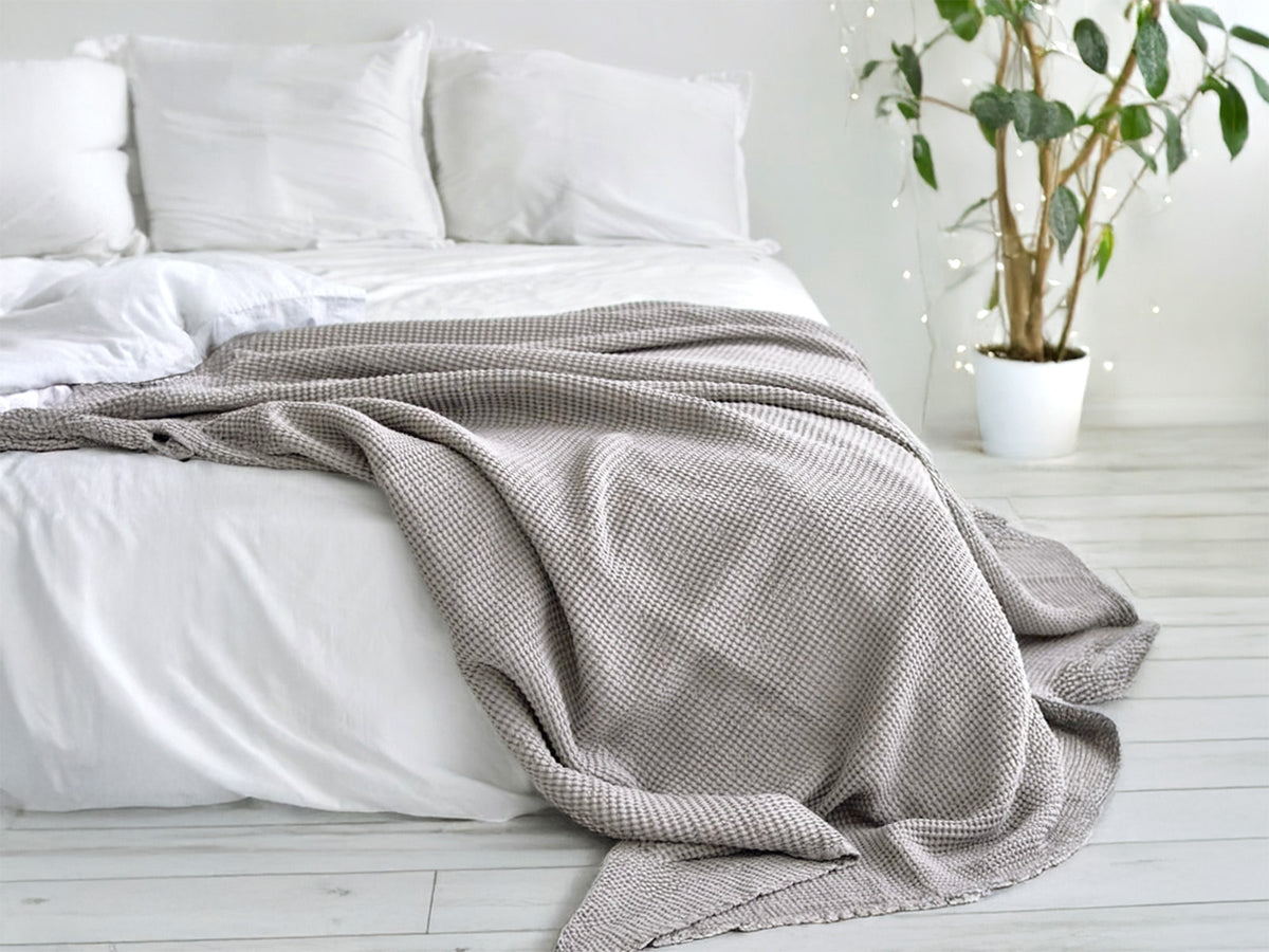 In einem Schlafzimmer liegt eine Nebel-graue Tagesdecke aus der beties "PUR.NATURE" Serie auf einem Bett ausgebreitet.